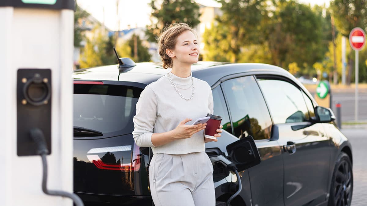 Woman uses a smartphone beside an electric vehicle connected to an outdoor charging station.